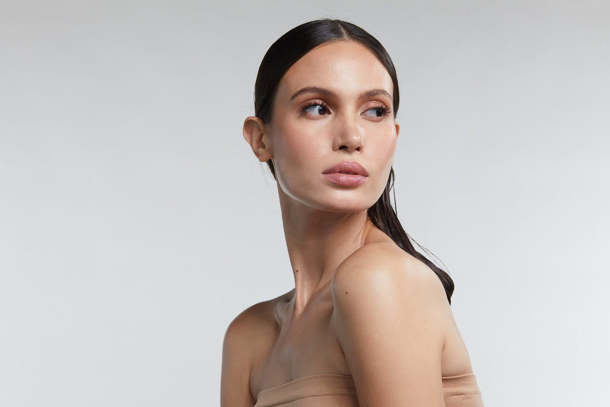 Close-up of a woman with radiant, clear skin and sleek hair looking to the side, representing the results of advanced skincare and LED light therapy treatments on a neutral background.