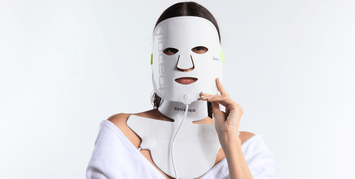 Woman wearing an LED light therapy face mask for acne treatment, in white robe against clean background, demonstrating at-home blue light therapy device for skin care