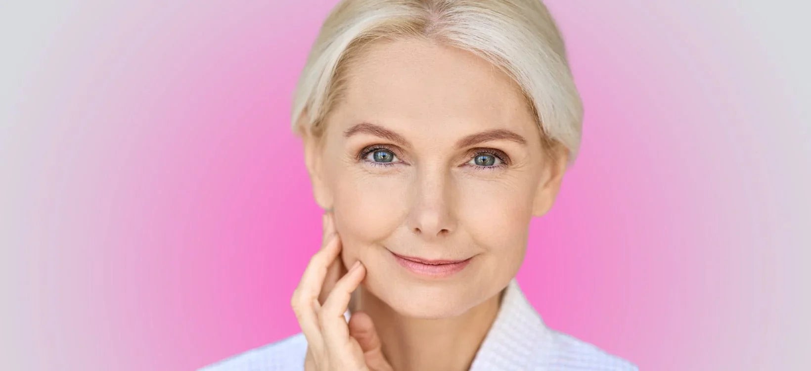 Smiling older woman with blonde hair, clear skin, and a gentle expression, touching her face with a soft pink gradient background.